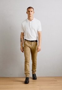 Hvit polo skjorte, khakibukser, svart belte og svarte sko. Modellen går på tregulv mot en ensfarget lys grå bakgrunn.