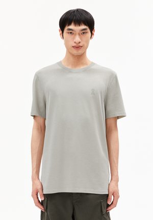 Mann in einem hellgrauen Kurzarm-T-Shirt mit kleinem Logo auf der linken Brust, kombiniert mit dunklen Cargo-Hosen.