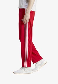 Rote Trainingshosen mit zwei weißen seitlichen Streifen, elastischem Bund und slim-fit Schnitt. Kombiniert mit weißen Sneakers und einem lockeren weißen Shirt.