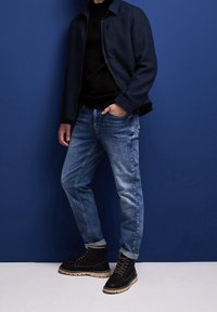 Mörkblå jacka, svart texturerad tröja, blekta blå jeans och svarta snörkängor med beige sula. Sittande mot en blå bakgrund.