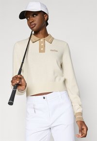 Γυναίκα που φορά λευκό καπέλο, μπεζ και κρεμ πουλόβερ πόλο, λευκό παντελόνι, κρατώντας ένα γκολφ μέταλλο που ακουμπάει στον ώμο της, μπροστά από ένα απλό φόντο.