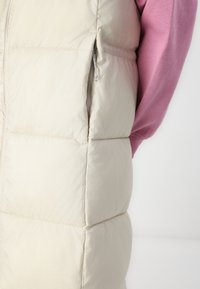 Lichtbeige puffer vest met een doorgestikte textuur, zijzak met rits, gedragen over een roze langemouwen kledingstuk.
