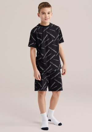 Băiat în picioare, purtând un tricou și pantaloni scurți negri asortați cu textul "NAP LEGEND" și șosete albe, pe un fundal neutru.