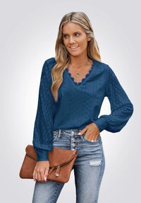 Marineblauwe blouse met gegolfde V-hals, gestructureerde mouwen, complexe borduursels, gecombineerd met lichtgekleurde jeans en een bruine clutch tas.