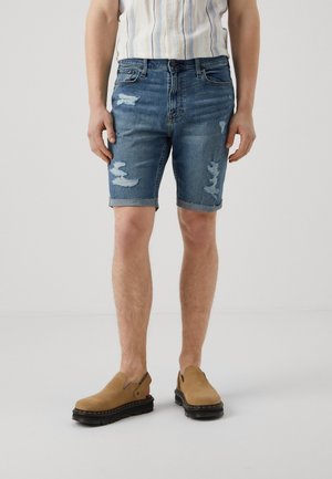 Person trägt blaue, ausgefranste Jeansshorts, beigefarbene Slipper und ein in die Hose gestecktes gestreiftes Hemd und steht vor einem einfarbigen Hintergrund.