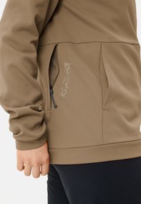 Hellbrauner Pullover aus strukturiertem Stoff, mit Reißverschluss-Seitentasche und Kordelzug. Mit bedrucktem Text in der Nähe der Tasche.