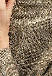 Tissu tissé dans des tons de brun et de gris avec un motif à carreaux. Le matériau semble texturé, mettant en valeur des détails et des coutures complexes.