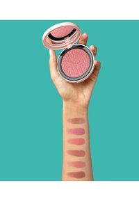 Kompaktes Rouge mit eingeprägtem Muster, pinke Farbe. Swatches am Arm zeigen sechs verschiedene Farbtöne von hellem bis dunklem Pink und Braun.