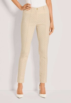 Frau trägt beige, eng anliegende Hose mit dezentem Diamantmuster und weißen High-Heels, steht mit einer Hand in der Hüfte.