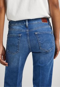 Blå denimjeans med mellanhöjd på midjan, femficksdesign och en läderpatch på bakdelen av midjan med kontrasterande söm.