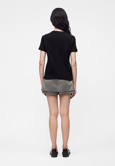 Femme aux longs cheveux foncés portant un t-shirt noir à manches courtes, un short gris superposé et des mocassins noirs, debout de dos sur un fond blanc uni.