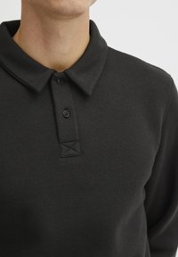 Svart polo-skjorte laget av mykt stoff, med kragehals, to-knaps knappestolpe og en etikett med X-stikking.
