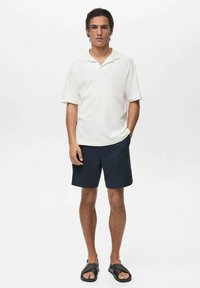 Polo shirt alb cu mânecă scurtă și guler plat, asortat cu pantaloni scurți închisi la culoare și sandale negre. Material neted și croi lejer.