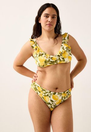 Pitkähiuksinen nainen, joka käyttää keltaista sitruunakuviollista bikiniä, jossa on röyhelöityjä olkaimia, seisoo käsi lantiolla.