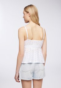 Top bianco in pizzo con spalline regolabili, design peplum e pattern ad occhiello. Abbinato a pantaloni corti in denim blu chiaro. Modella girata di spalle.