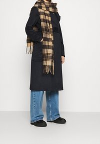 Persoon in een lange zwarte jas, blauwe jeans, zwarte schoenen en een grote beige en bruine geruite sjaal tegen een effen witte achtergrond.