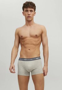 Νεαρός άντρας με κοντά σπαστά μαλλιά, φορώντας ανοιχτό γκρι μποξεράκι με λογότυπο στη ζώνη, στέκεται μπροστά σε ένα απλό ανοιχτό φόντο.