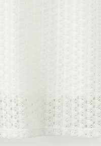 Tissu blanc avec un motif de dentelle complexe comprenant des boucles répétées et des motifs floraux, créant une apparence délicate et texturée.