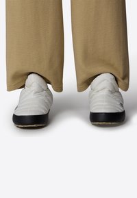 Bílé prošívané slip-on boty s černou gumovou podrážkou, částečně zakryté béžovými proužkovanými kalhotami, které ukazují hladkou texturu a zaoblený design.