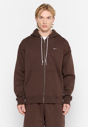 Brązowa bluza z kapturem zapinana na zamek, wykonana z miękkiego materiału, z kieszenią typu kangur oraz białymi sznurkami, z małym logo Nike na piersi.