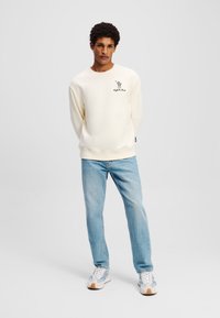 Kreminės spalvos crewneck džemperis su juodu logotipu, derinamas su šviesiai mėlynomis džinsinėmis kelnėmis. Modelis vilki baltus ir mėlynus sportbačius, stovintis neutraliame fone.