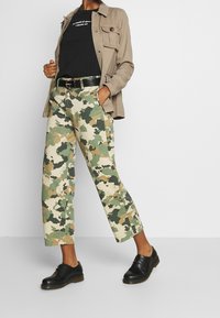 Pantalones con estampado de camuflaje en verde y beige, combinados con una camiseta gráfica negra y una chaqueta de botones en color marrón claro. Zapatos negros con suela resistente.