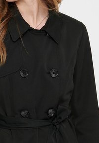 Schwarzer Trenchcoat aus glattem Stoff, mit einem doppelt geknöpften Design, großen Knöpfen, Reverskragen und einem gebundenen Taillengürtel.