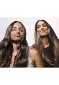 Deux femmes aux longs cheveux ondulés, foncés. L'une porte un débardeur clair, et l'autre une chemise sombre. Toutes deux ont une peau lisse sur un fond lumineux.