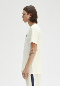 Camiseta de manga corta color crema, con cuello redondo, que presenta un pequeño logo negro en el pecho. Combinada con pantalones de color claro. Textura suave.