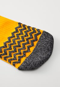 Gul strumpa med ett svart zigzag-mönster upptill och en mörkgrå tå. Texturen verkar vara stickad med en slät yta.
