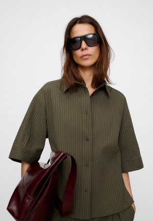 Frau mit schulterlangem braunem Haar, trägt schwarze Sonnenbrille, ein grünes Nadelstreifenhemd und hat eine große dunkelrote Ledertasche.