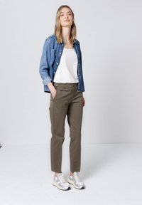 Femme debout portant une chemise en jean bleue par-dessus un haut blanc, un pantalon vert olive avec des poches latérales, et des baskets argentées et blanches sur un fond uni.