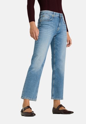 Personne portant un jean droit bleu clair, un haut à manches longues marron foncé et des chaussures plates pointues marron foncé avec boucles.