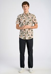 Korte mouwen overhemd met knopen in lichtbeige met een bloemen- en bladerenpatroon; gecombineerd met zwarte broeken en witte sneakers.