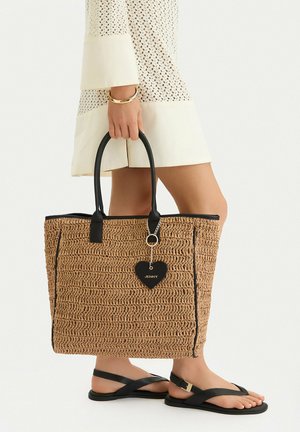 Žena v bílých pletených šatech držící velkou tkanou světle hnědou tašku s černými úchyty a přívěskem ve tvaru srdce, obutá v černých sandálech.