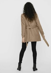 Beige trenchcoat met riem, enkelvoudige knoopsluiting, lange mouwen met opgerolde manchetten, gecombineerd met zwarte skinny jeans en haklaarzen.