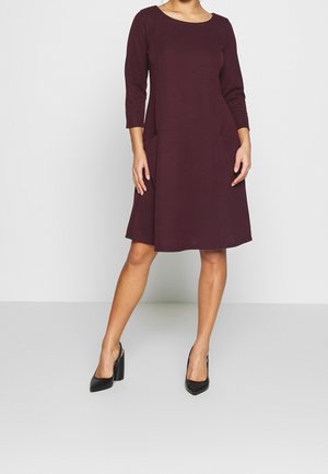 Mujer con vestido morado oscuro hasta la rodilla, con mangas tres cuartos y zapatos de tacón negro, de pie frente a un fondo liso.