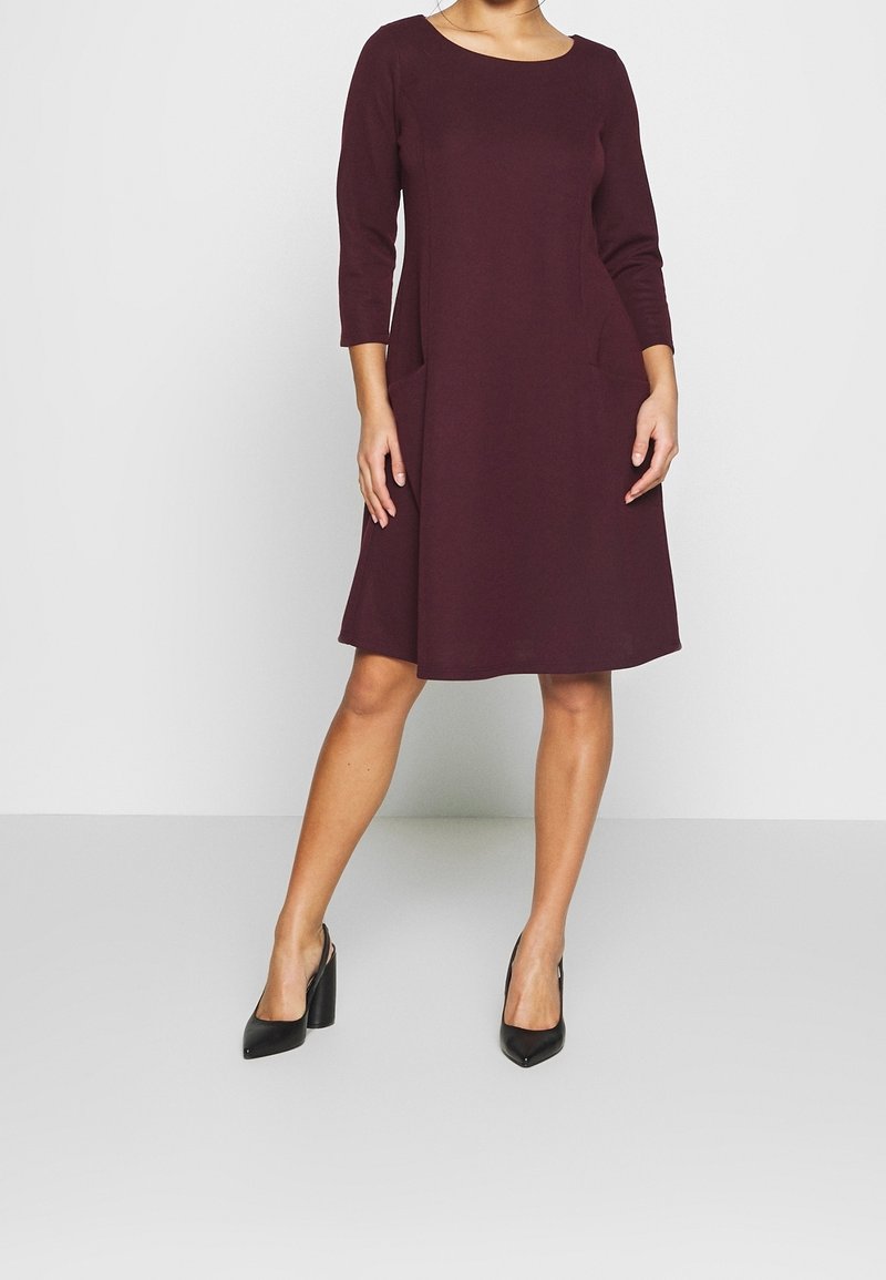 Femme portant une robe violette foncée au genou avec des manches trois-quarts et des chaussures à talons noirs, se tenant devant un fond uni.