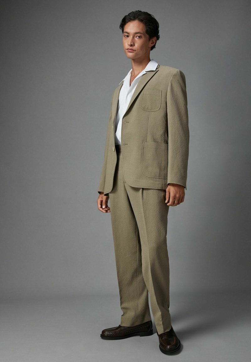 Light brown textured suit with large pockets and a collared white shirt, complementing dark shoes, displayed on a gray background.