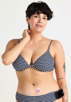 Niebiesko-białe pasiaste bikini z regulowanymi ramiączkami, z trójkątnym biustonoszem i pasującym dołem; na brzuchu widoczny jest aparat medyczny.