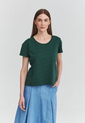 Vrouw met lang bruin haar draagt een donkergroen T-shirt met korte mouwen en een lichtblauwe geplooide rok, staand tegen een effen achtergrond.