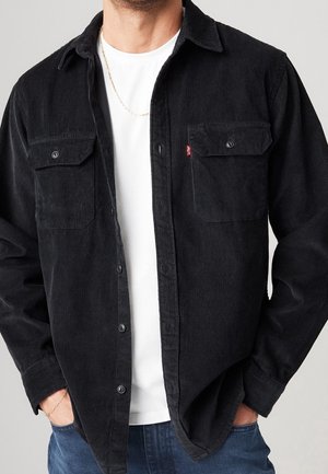 Homme portant une chemise en velours côtelé noire boutonnée sur un T-shirt blanc, un jean bleu, une chaîne en or au cou et un bracelet, les mains dans les poches.