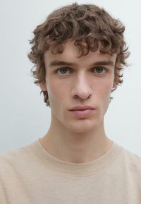 Primo piano di un uomo con capelli castani corti e ricci e pelle chiara, che indossa una maglietta beige chiaro con scollo rotondo su uno sfondo neutro.