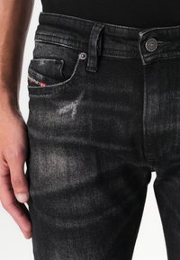 Black denim jeans with a faded, worn texture. Features include a front pocket, metal button, and a visible brand tag in red.