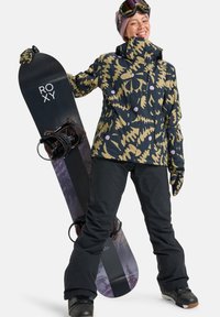 Snowboard mit lila und schwarzem Farbverlauf, gehalten von einer Person in einer gemusterten Jacke, schwarzen Hosen und Snowboard-Goggles.