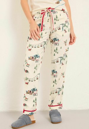 Hands resting on waist, wearing cream pajama pants with ski lodge and cable car print and gray slip-on slippers with brown soles.