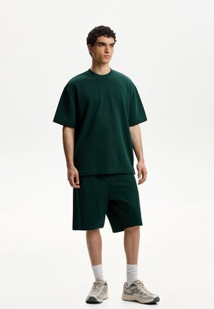 Dark green cotton t-shirt with short sleeves, paired with matching shorts. Model wears white socks and light gray sneakers. Simple design.