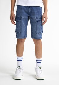 Džínové cargo šortky ve středně modré barvě s dvěma velkými bočními kapsami, roztrhaným lemem a volným střihem. Spárovány s bílými teniskami a modro-proužkovanými ponožkami.