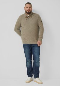 Beige gebreide trui met een gestructureerd patroon, sjaalkraag en toggles, gecombineerd met een blauwe spijkerbroek en witte sneakers.