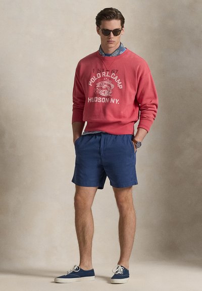 Sweatshirt rouge avec logo blanc, short bleu marine et baskets bleues. Le modèle porte des lunettes de soleil et un bandana bleu à motifs autour du cou.